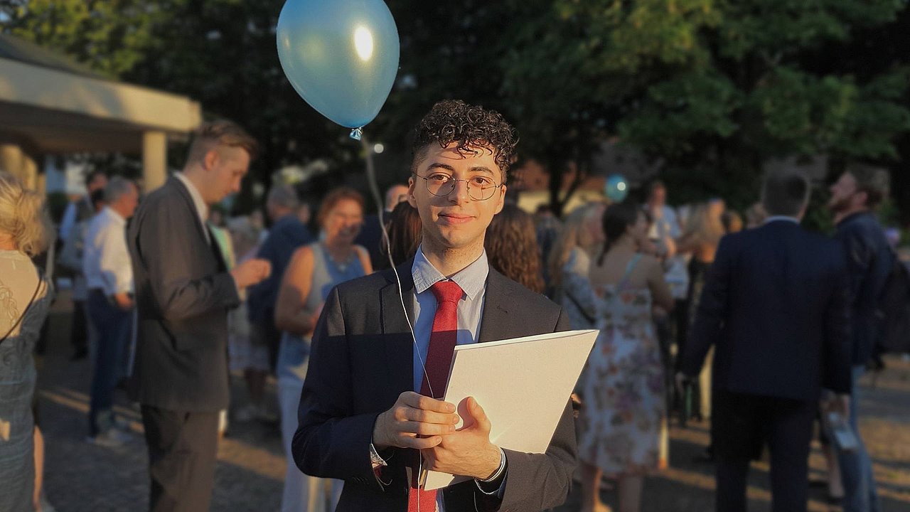 Deeb steht in Menschenmenge mit Abizeugnis in der Hand und Luftballons