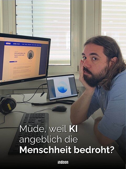 Ein Mann mit langen braunen Haaren und blauem Hemd sitzt an einem Arbeitsplatz vor mehreren Bildschirmen. Er blickt müde in die Kamera. Text: Müde, weil KI angeblich die Menschheit bedroht?