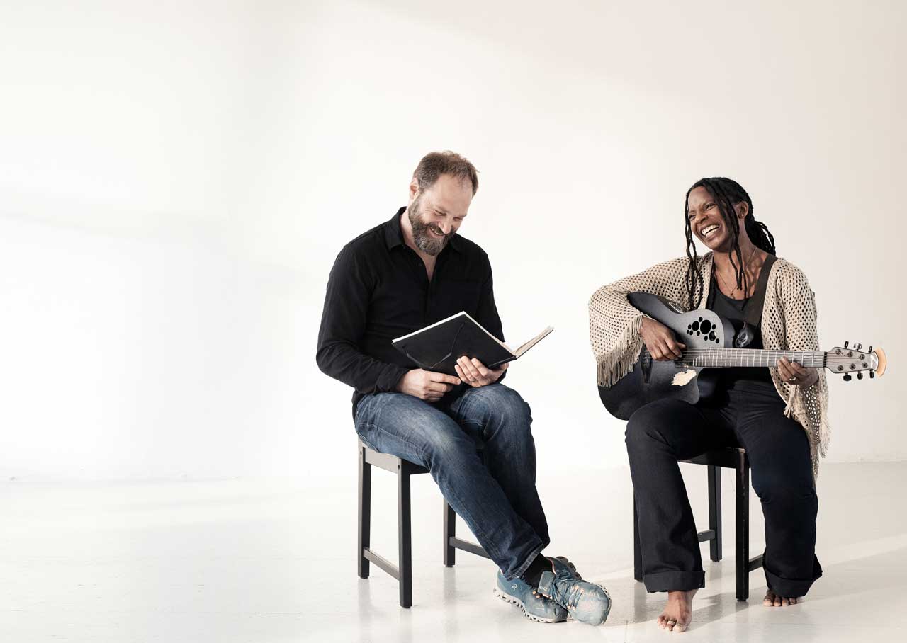 Patrick und Judy sitzen auf Stühlen im Fotostudio. Er hält ein Buch und sie ihre Gitarre. Beide lachen. 