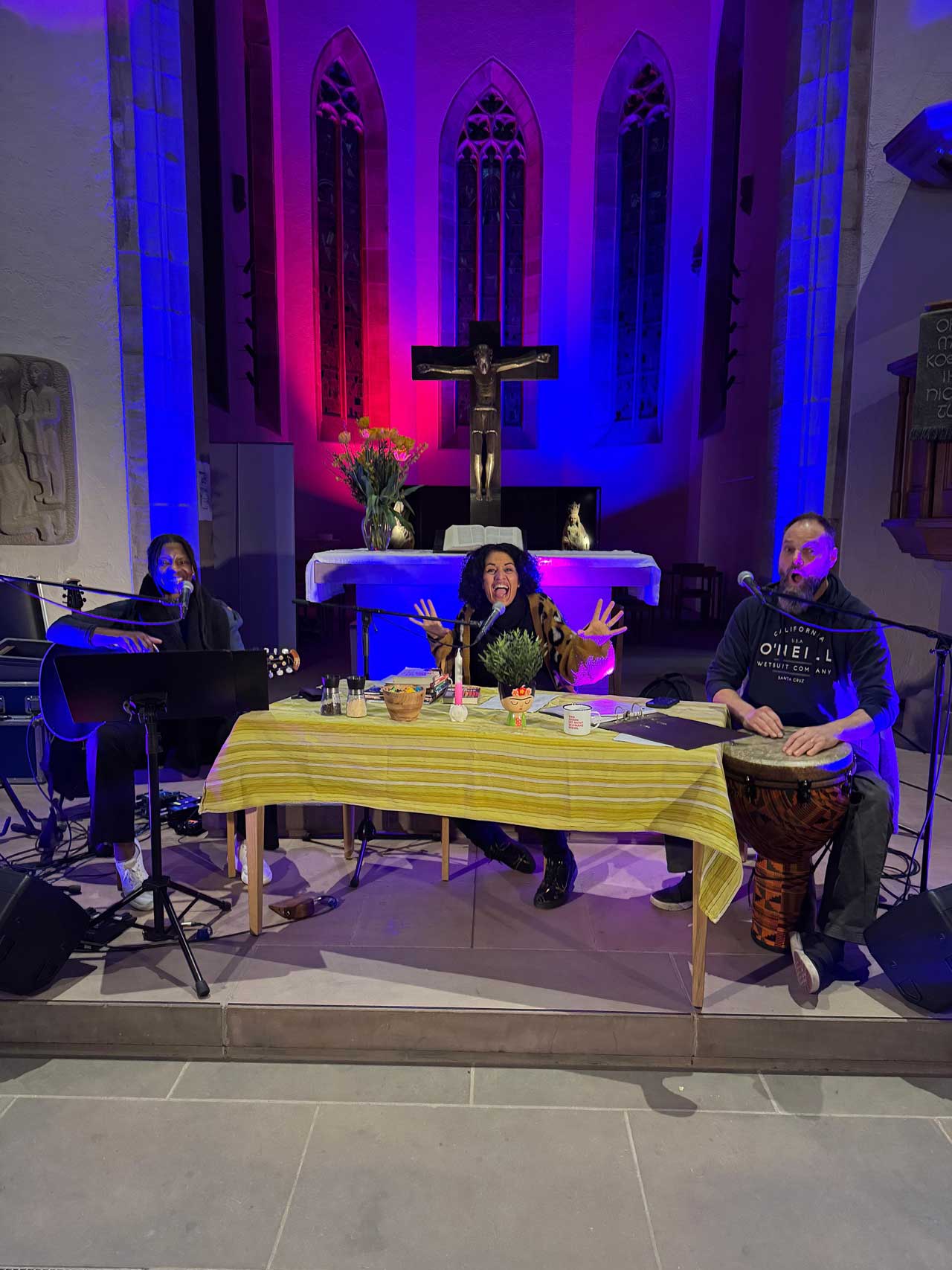 Konzertlesung in einer Kirche mit Judy Bailey & Patrick Depuhl. Links ist Judy an der Gitarre, in der Mitte Sarah und rechts Patrick an einer Trommel.