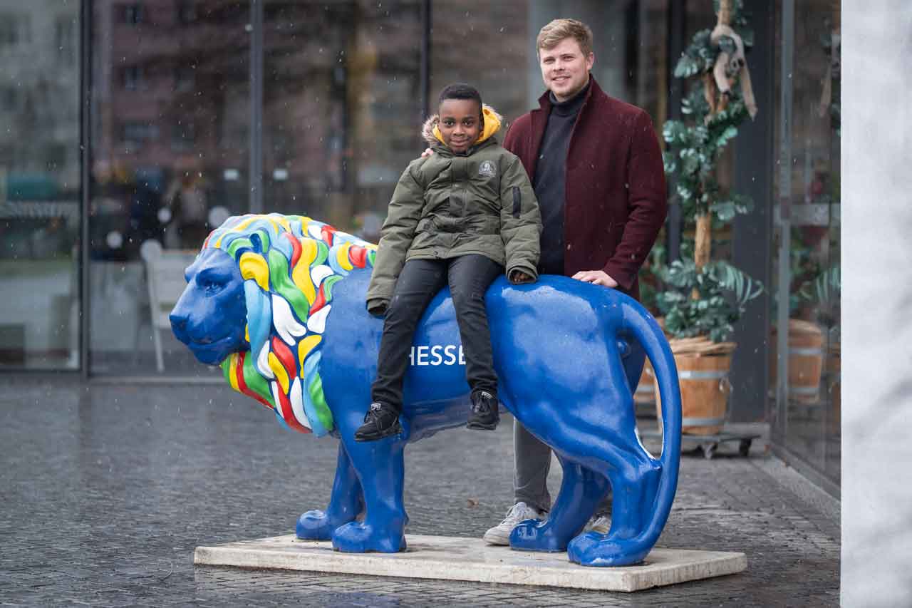 Richmond sitzt auf einem künstlichen blauen Löwen vor dem Eingang des Frankfurter Zoos. Mathias steht hinter ihm.