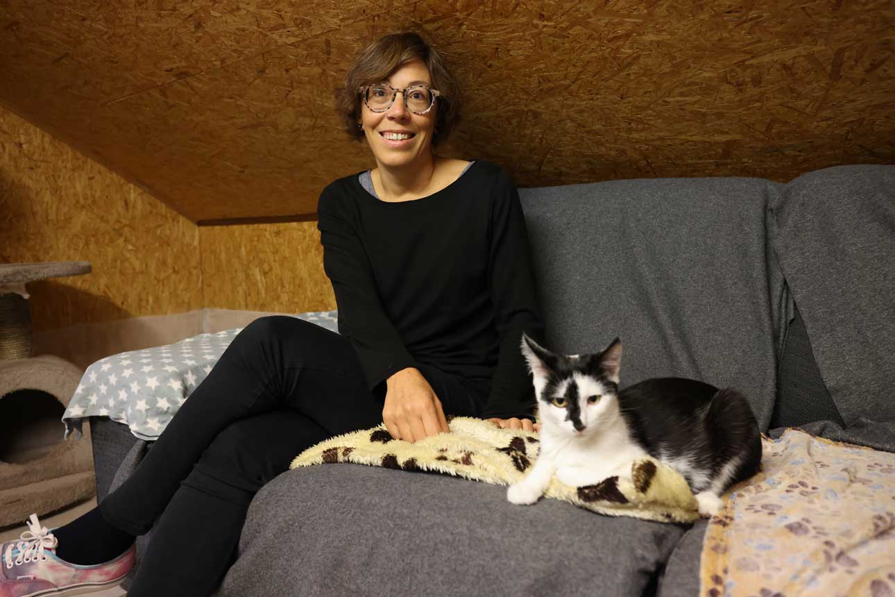 Marlene sitzt mit einer schwarz-weiß gestreiften Katze auf der Couch und lächelt in die Kamera.