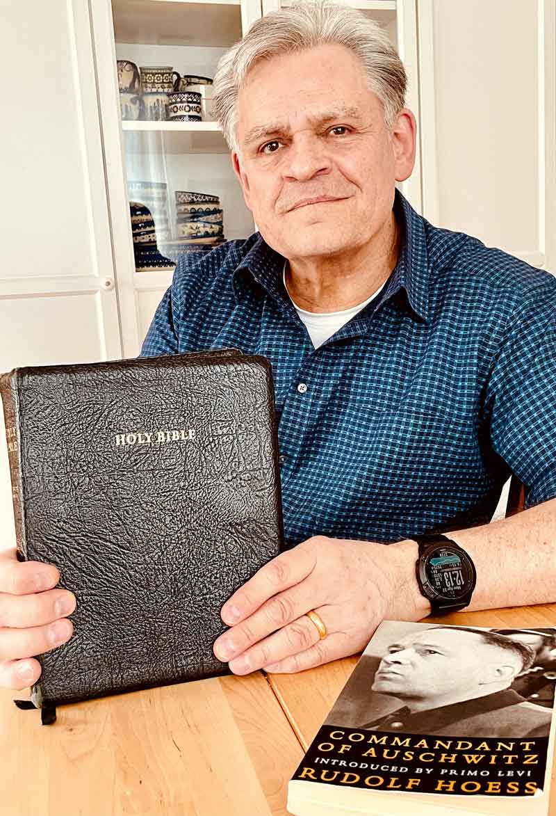 Kai Uwe Höss sitzt am Tisch und hält eine Bibel in der Hand. Daneben liegt ein Buch mit dem englischen Titel „Commandant of Auschwitz Rudolf Hoess“ .