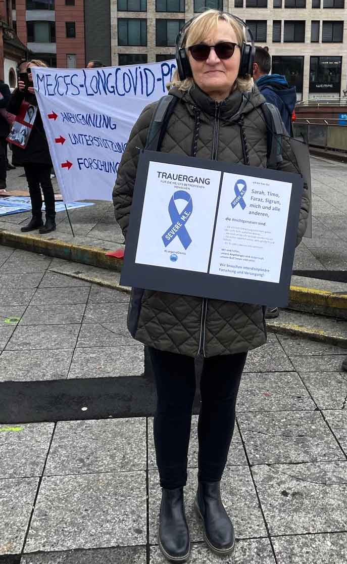 Renate steht mit einem Schild an der Frankfurter Hauptwache. Darauf steht Trauergang. Im Hintergrund ist ein Banner mit der Aufschrift: ME/CFS-LongCovid - Anerkennung, Unterstützung, Forschung