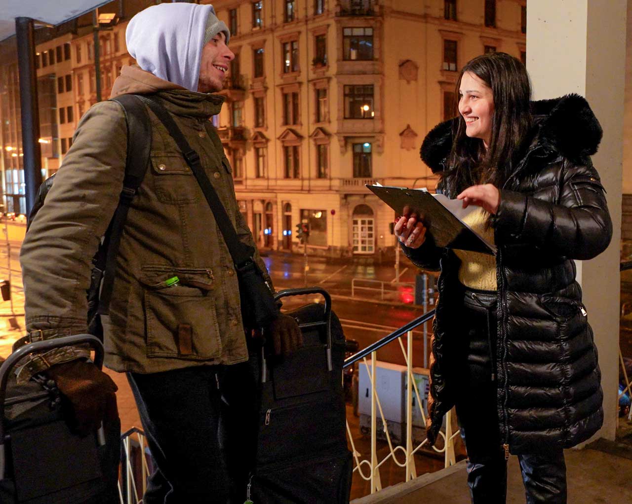 Geania mit ihrer Liste vor der Tür der Weißfrauen Diakoniekirche. Neben ihr lächelt sie ein junger Mann an. Er hat 2 Taschen dabei.