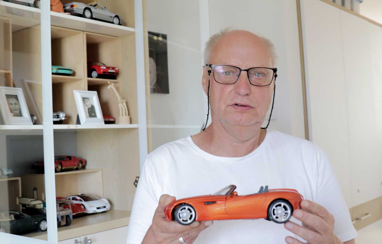 Gernot zeigt ein orangefarbenes Modellauto. Im Regal hinter ihm stehen weitere Modellautos und Fotos.