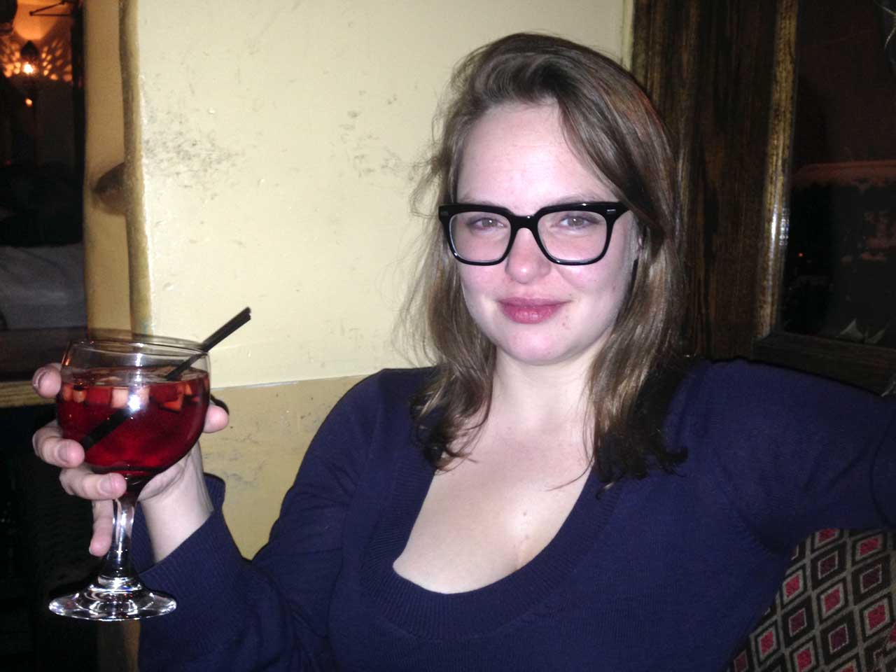 Nathalie hält ein Weinglas mit einer roten Flüssigkeit in der Hand. Es könnte Sangria sein. Sie blickt entspannt in die Kamera. 