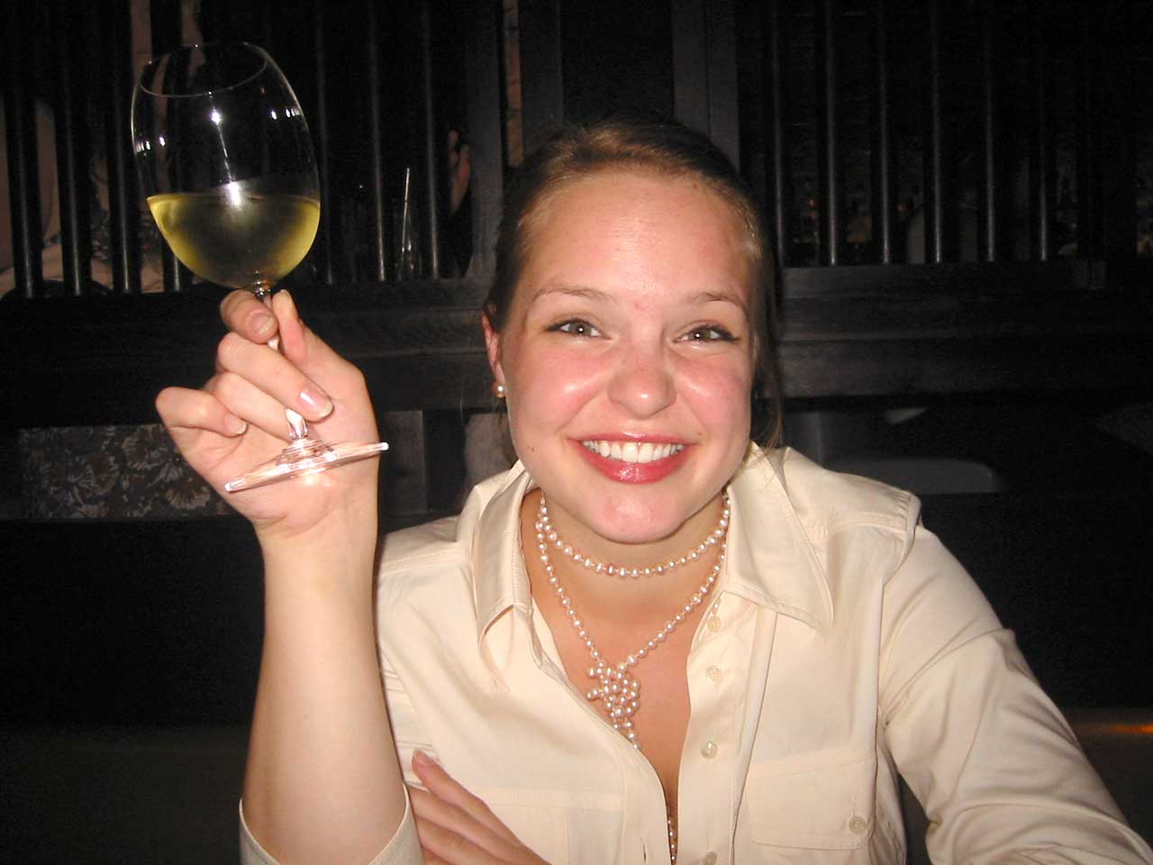 Nathalie hält ein Glas Weißwein in der Hand. Sie lächelt in die Kamera. 