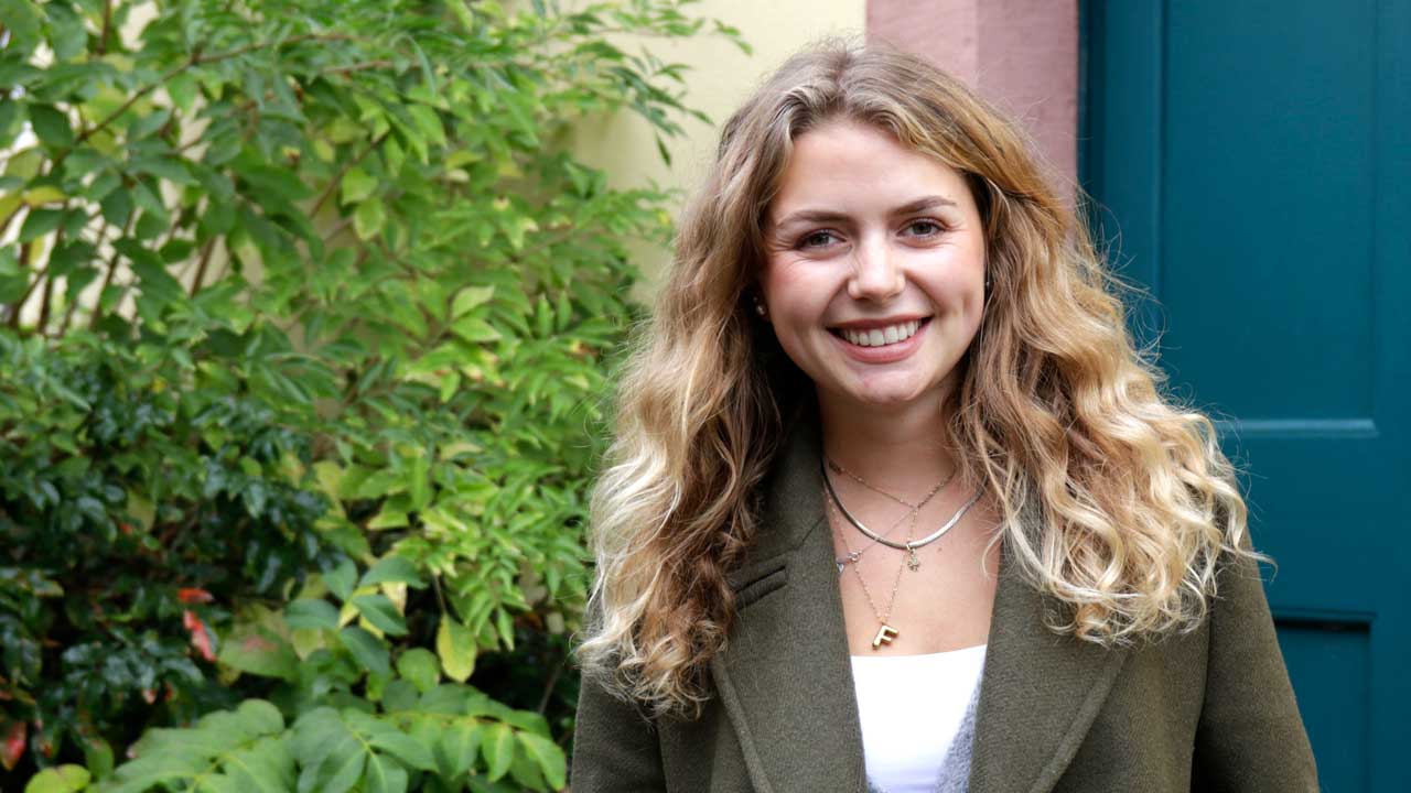 Floriane steht draußen vor einem grünen Strauch. Die blonde Frau lächelt in die Kamera.