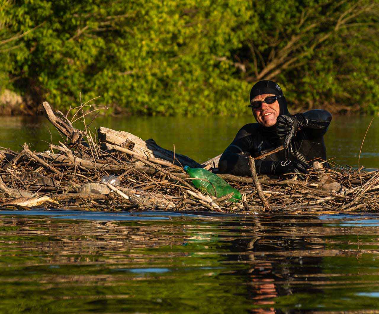 Schwimmendes Gehölz in einem Fluss. Darin mehrere Plastik-Flaschen. Andreas Fath ist hingeschwommen und blickt in die Kamera. Der Daumen zeigt nach unten.