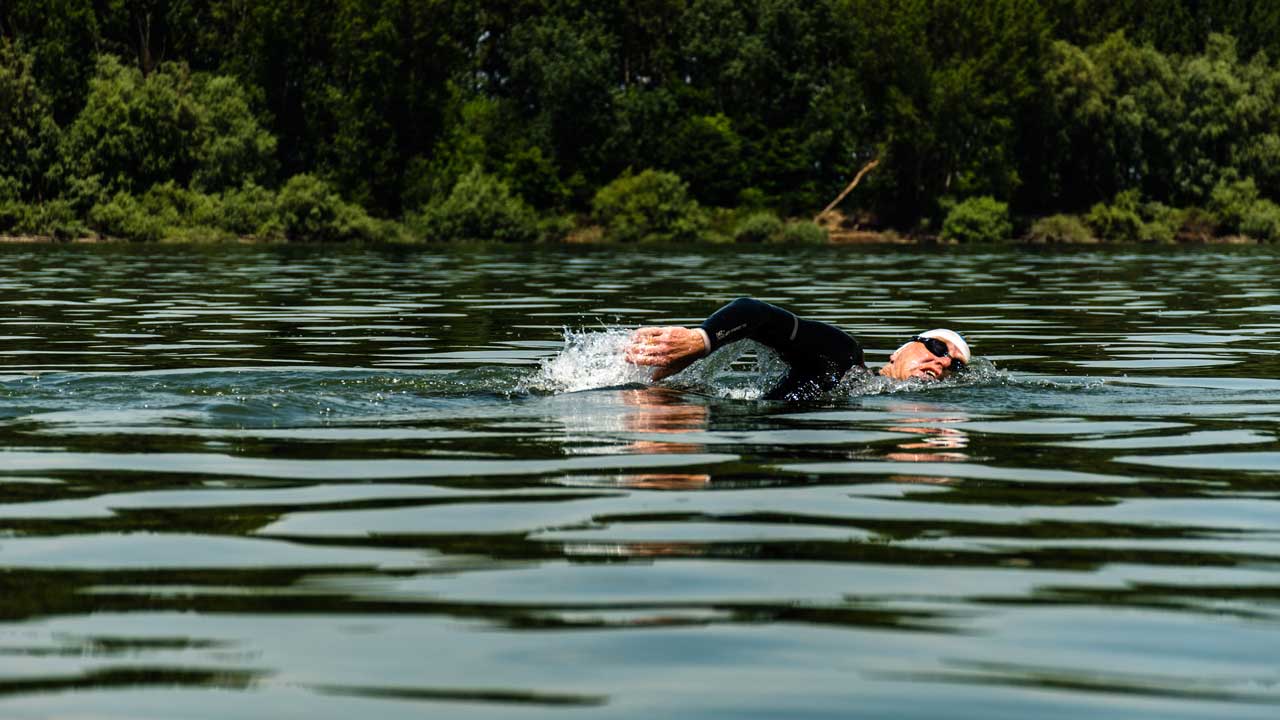 Schwimmend im Fluss. Andreas krault und holt gerade Luft. Im Hintergrund sieht man das Ufer.