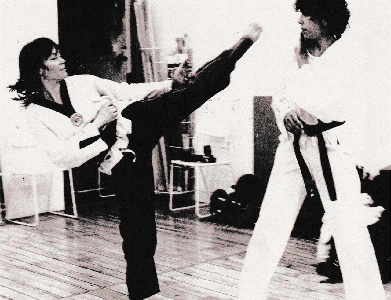 Man sieht auf dieser alten aufnahme Sunny Graff und die Mitgründerin von "Frauen in bewegung". Die beiden Fraueen tragen Taekwondo-Anzüge und trainieren gegen einander. Sunny trittt der anderen Frau dabei auf Kopfhöhe. 