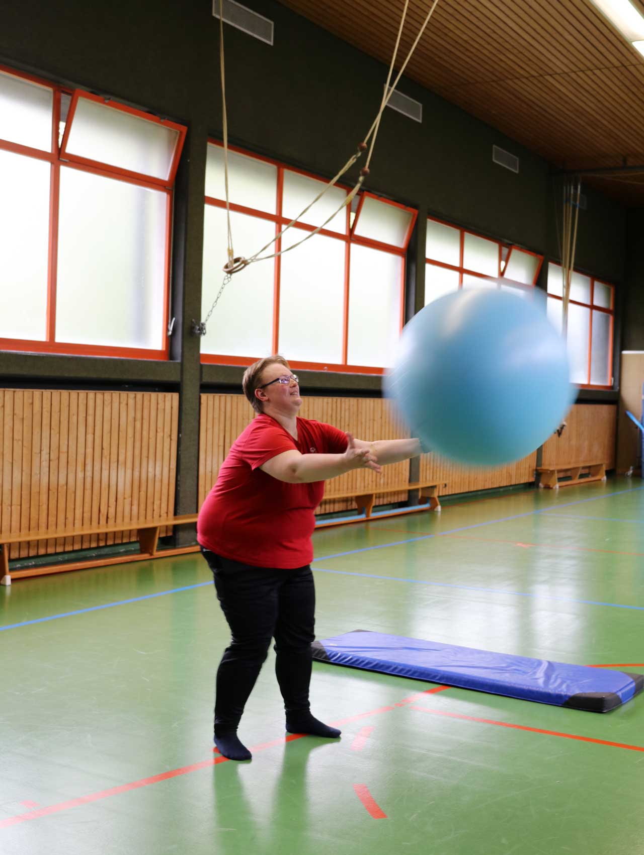 Sabrina wirft einen großen Ball jemandem zu. Sie ist in einer Turnhalle.