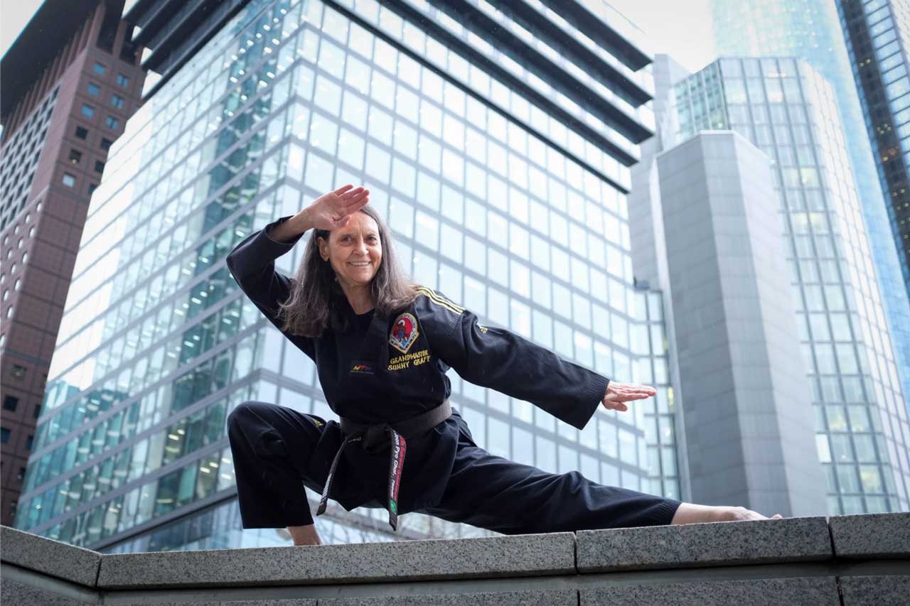 Die Takwondo-trainerin Sunny graff posioert in einer Taekwondo-position vor Hochhäusern, Sie trägt einen schwarzen Taekwondo-Anzug und lächelt.  