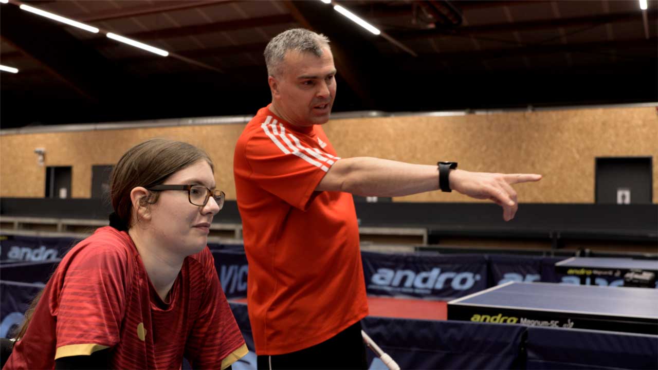 Jana und ihr Trainer in der Halle - er zeigt nach vorne auf die andere Seite der Platte, an der sie stehen.