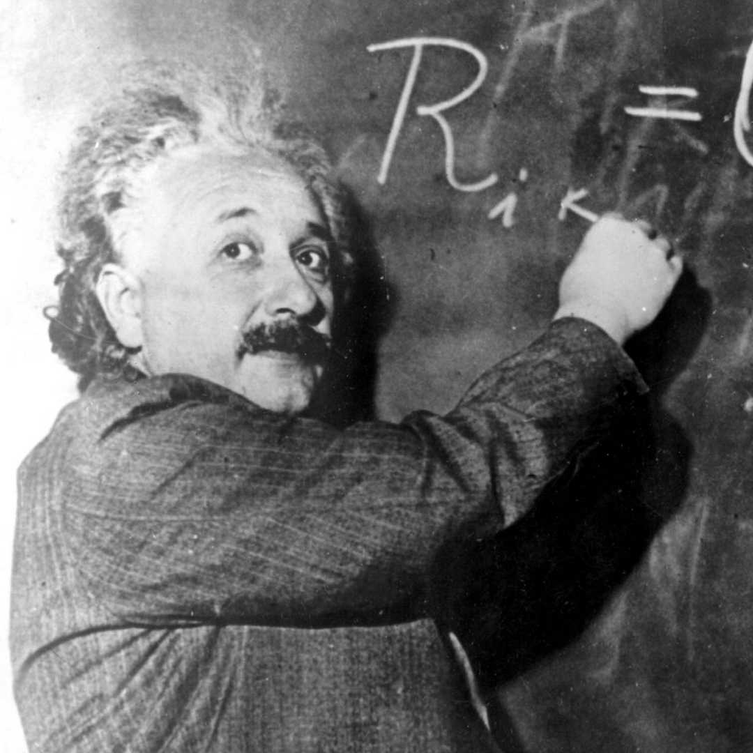 Das Schwarz-Weiß-Foto zeigt den Mathematiker Albert Einstein vor einer Tafel. Er blickt in die Kamera und ist gerade dabei etwas mit Kreide auf die Tafel zu schreiben