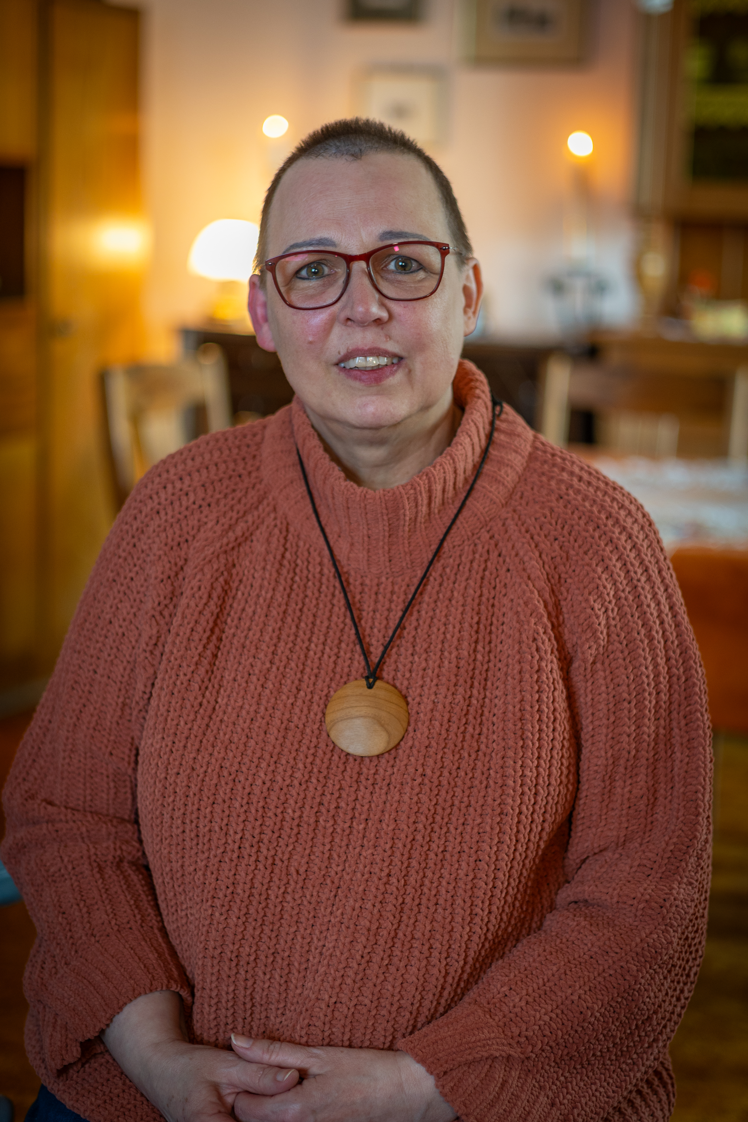 Maike Kniese mit roter Brille und rosafarbenem Strickpullover sitzt in einem warm beleuchteten Wohnzimmer.
