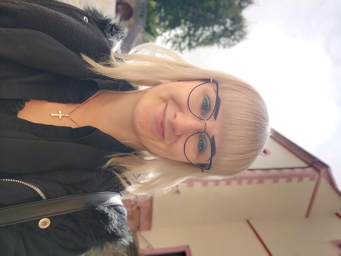Zu sehen ist die Vikarin und Finnin Jenni Kempcke. Sie steht vor einem Gebäude. Es handelt sich um eine Selfieaufnahme. Jenni Kempcke hat blonde Haare und trägt eine Brille. Sie trägt eine schwarze Bluse und eine schwarze Jacke darüber, welche leicht geöffnet ist.