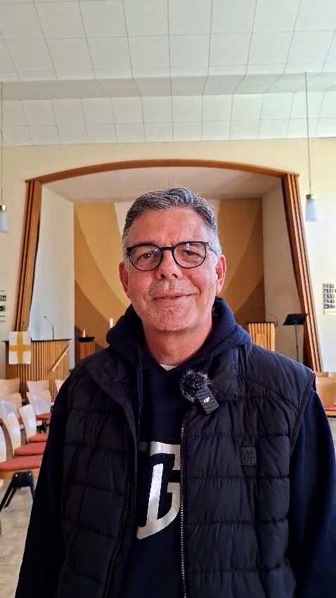 Jan Spangenberg steht  mit Brille und dunkler Weste lächelnd in einer modernen Kirche.