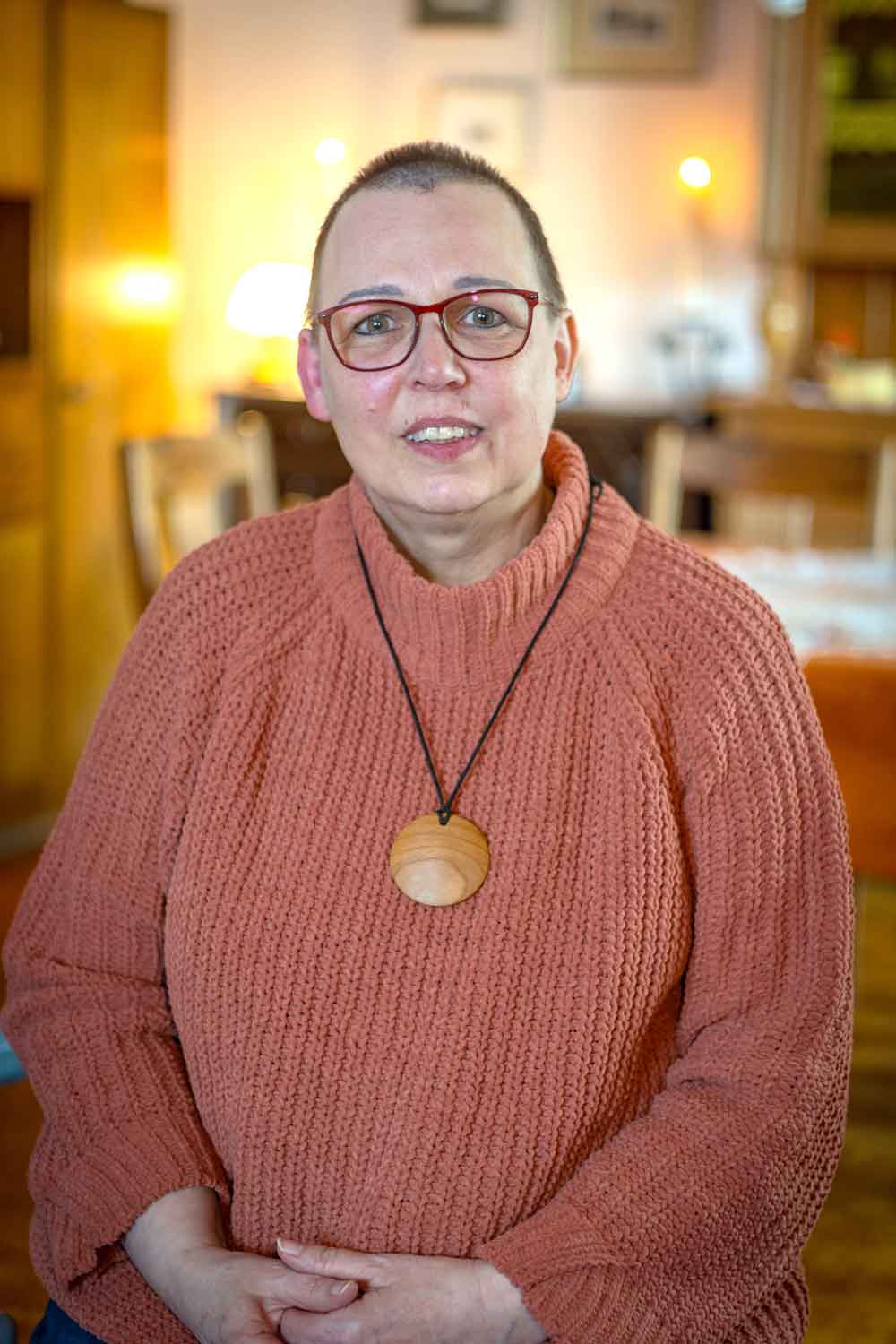 Maike Kniese mit roter Brille und rosafarbenem Strickpullover sitzt in einem warm beleuchteten Wohnzimmer.