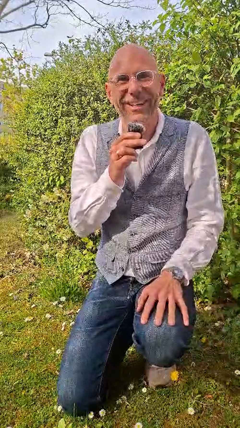 Martin Vorländer, ein lächelnder Mann mit Brille, kniet in einem blühenden Garten und hält ein Mikrofon.
