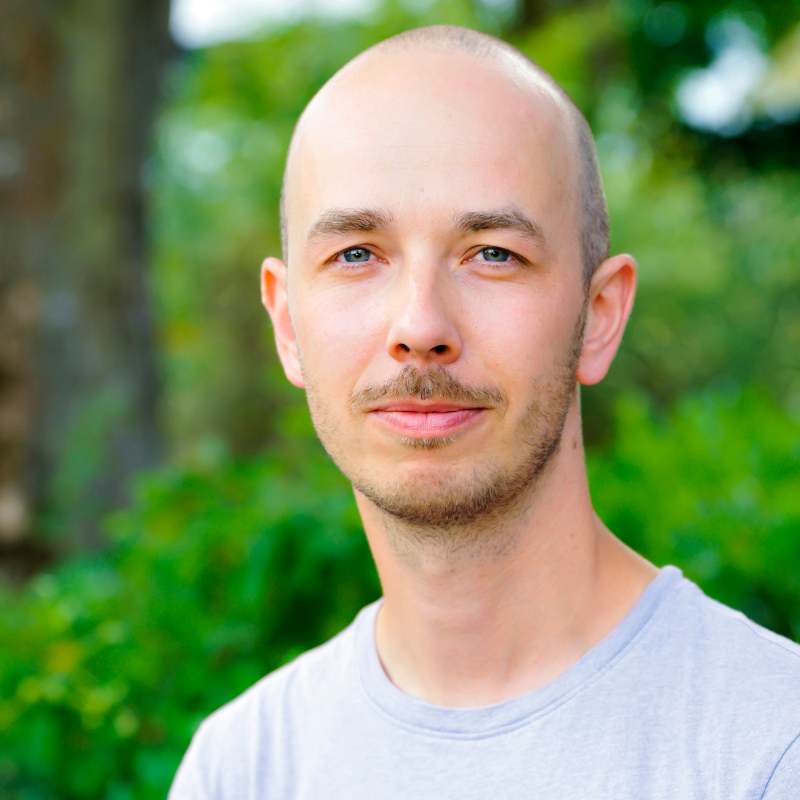 Neutral lachender Mann mit Glatze blickt in die Kamera. Er trägt ein hellblaues Shirt.