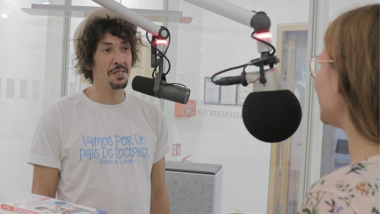 Danny Beuerbach steht am Mikro im gläsernen Studio und schaut Charlotte Mattes an. 