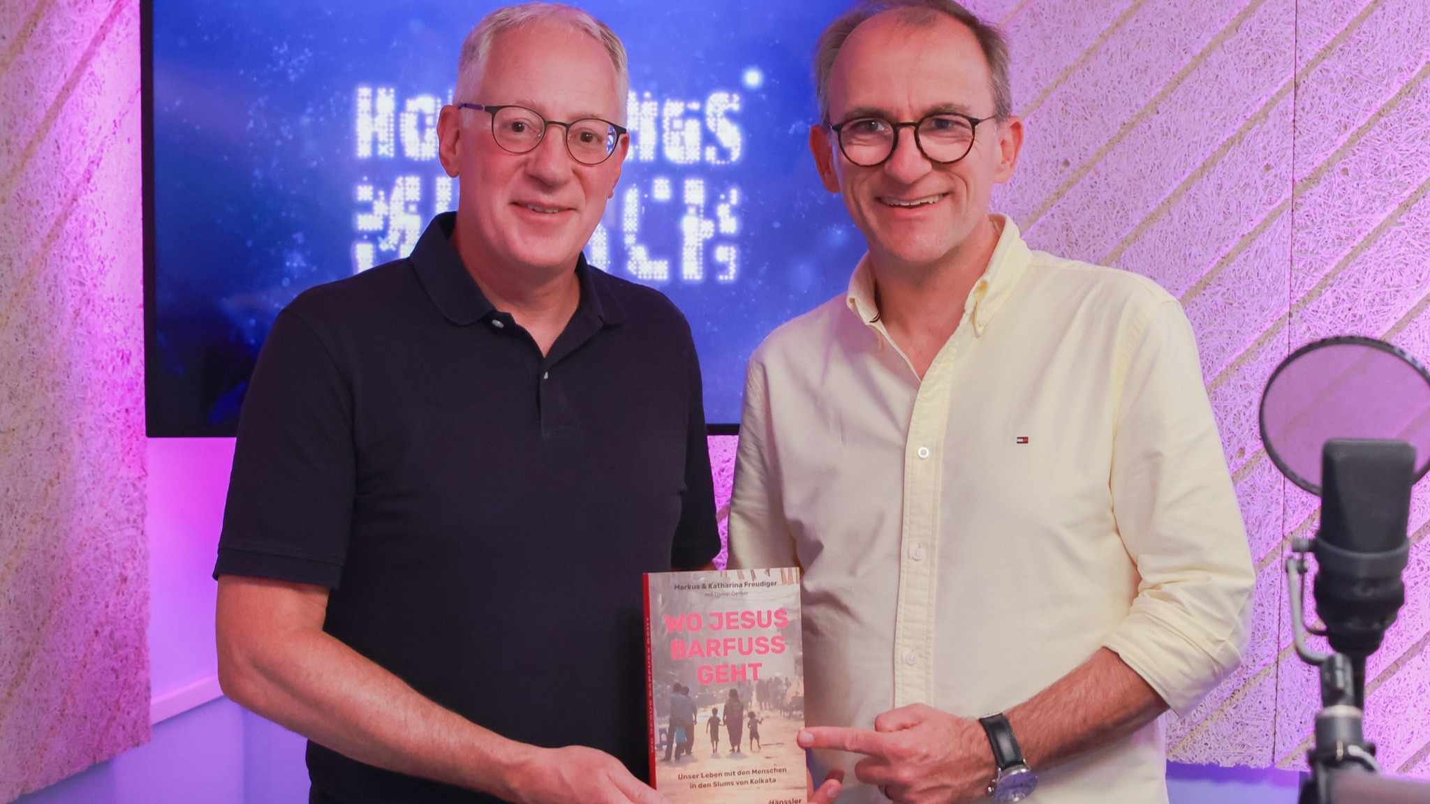 Ein Mann mit schwarzem Shirt, weißen Haaren und Brille hält ein Buch. Daneben ein zweiter Mann mit weißem Hemd, Brille und grauen Haaren. Podcastmikrofon daneben. Im Hintergrund pinke Wand und blauer Screen mit Aufschrift "Hoffnungsmensch".