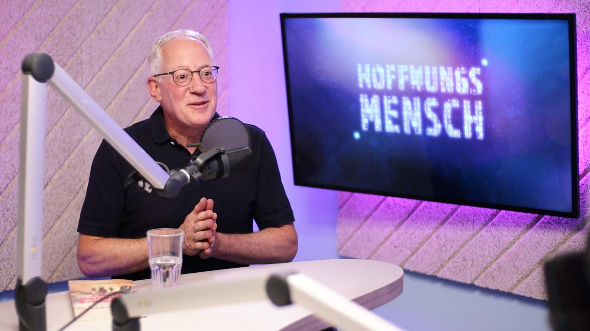 Ein Mann mit schwarzem Shirt, weißen Haaren und Brille hinter Podcastmikrofon. Im Hintergrund pinke Wand und blauer Screen mit Aufschrift "Hoffnungsmensch".