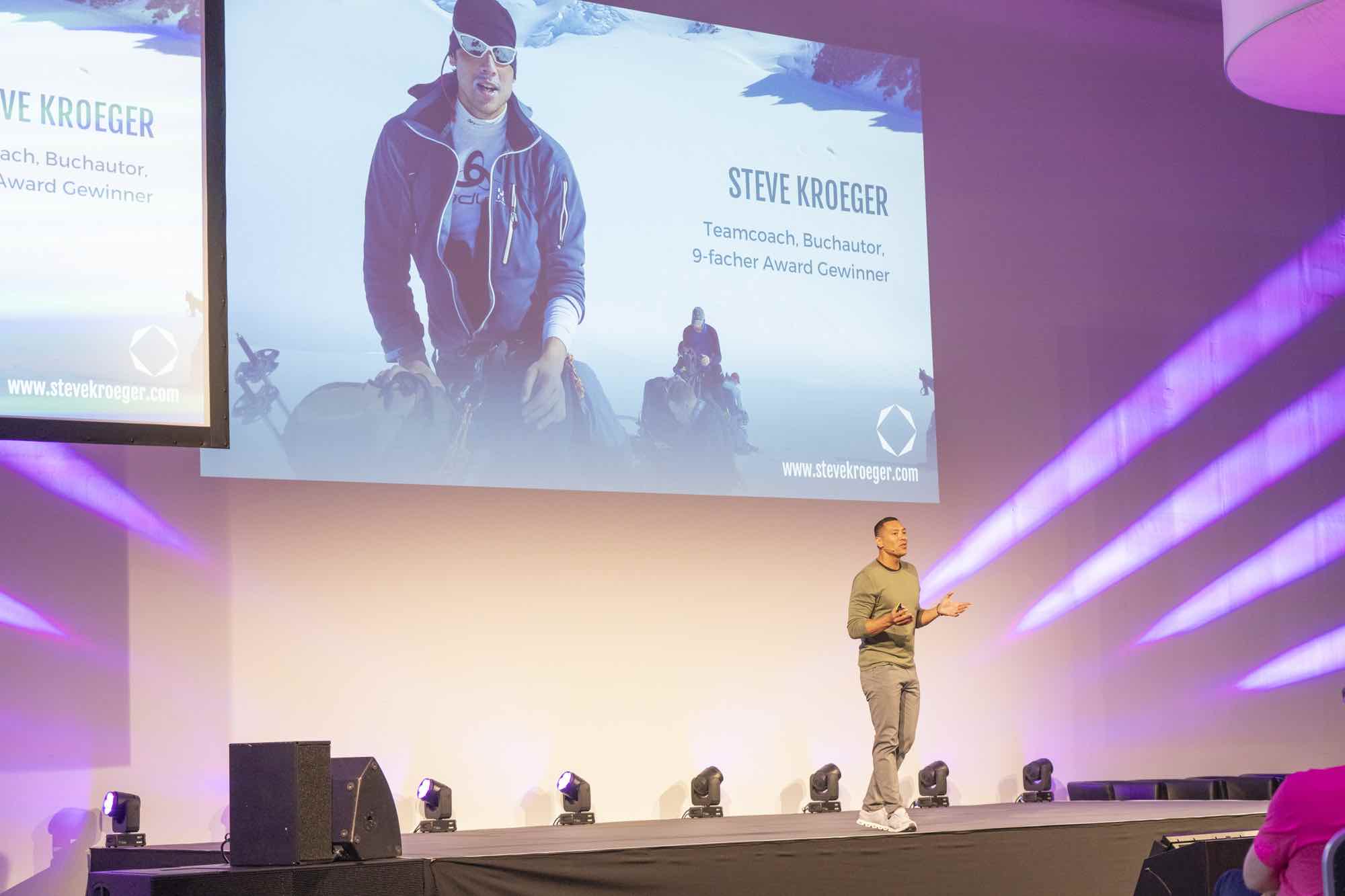 Das Bild zeigt Steve auf einer Bühne. Er hält gerade einen Vortrag. Auf der Leinwand im Hintergrund ist ein Bildd von ihm zu sehen, dass ihn auf einer seiner Bergsteigertouren im Schnee zeigt.