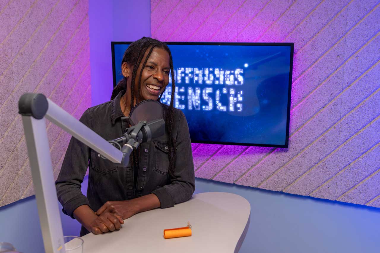 Judy steht am Mikrofon im Tonstudio und lächelt den Host an. Steffen Kern ist nicht im Bild. Im Hintergrund ist ein großer Bildschirm, auf dem steht: HOFFNUNGSMENSCH
