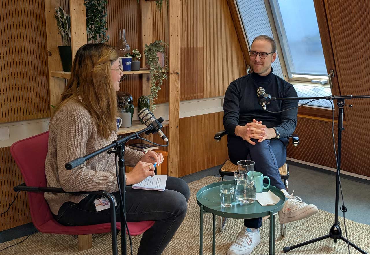 Host Charlotte und Lukas Freund sitzen sich gegenüber im Studio. Sie lächeln sich an und gucken nicht in die Kamera. 