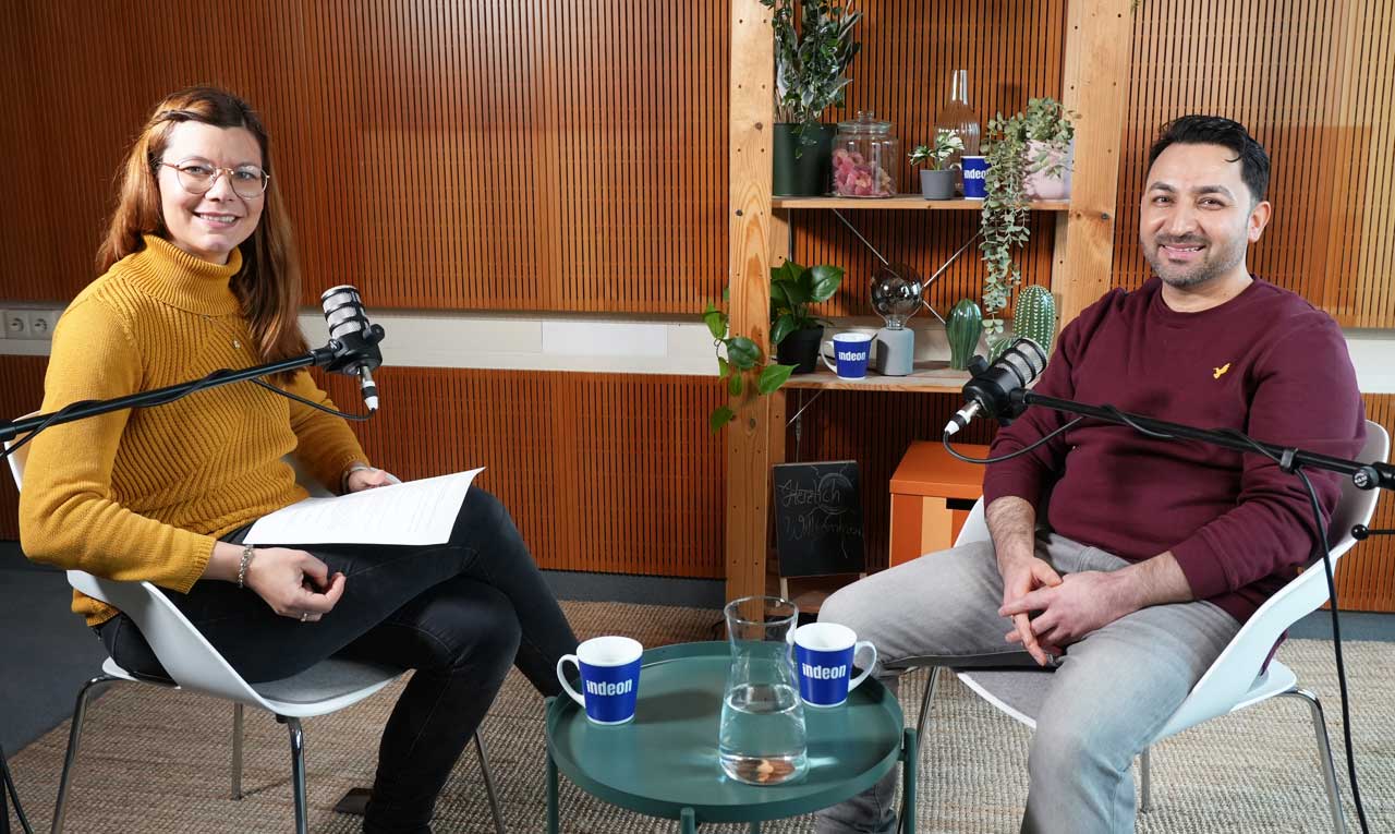 Host Charlotte Mattes, in gelbem Pulli, schaut in die Kamera. Daneben steht ein kleiner Tisch mit Indeon-Tassen darauf. Parwiz, die Hände im Schoß gefaltet, lächelt auch in die Kamera. 