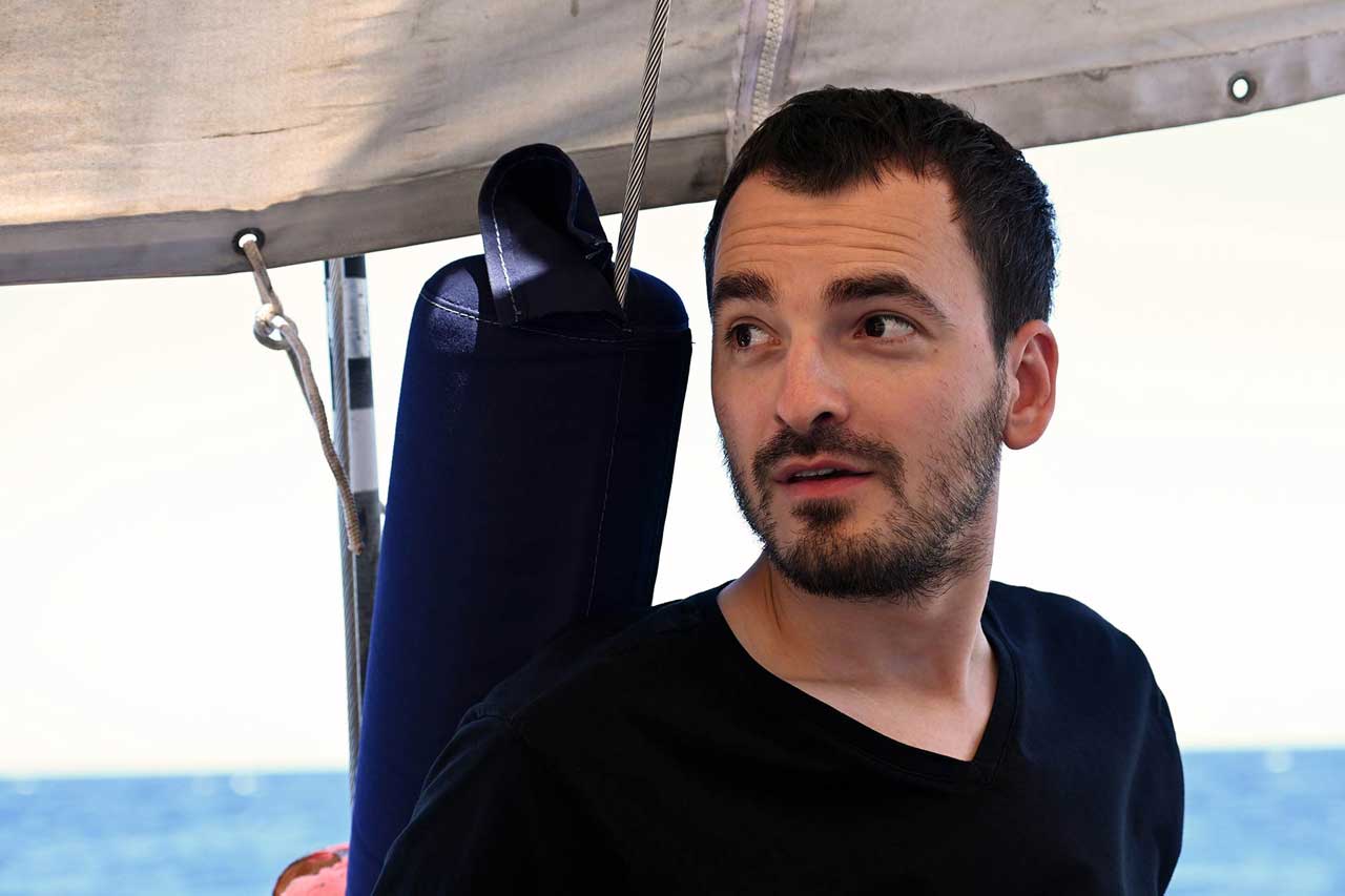 Stefen ist auf Schiff, guckt links an der Kamera vorbei. Über ihm ein Segel. Im Hintergrund das Mittelmeer. 