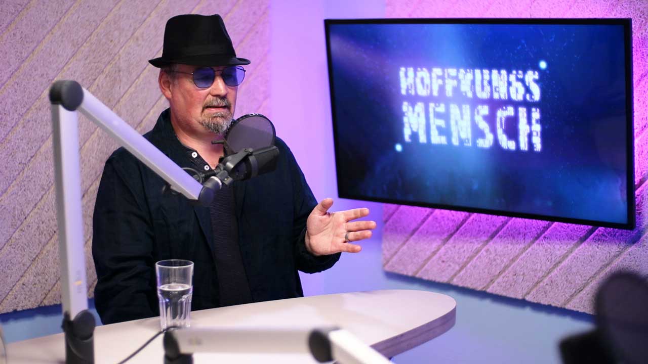 Leo im Podcast-Studio. Er trägt eine blaue Brille und einen schwarzen Hut. Im Hintergrund ein Bildschirm mit dem Hoffnungsmensch-Logo. 