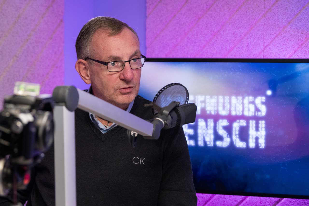 Bernd Siggelow im Tonstudio. Vor seinem Mund ein Mikrofon im Hintergrund ein Fernseher auf dem „Hoffnungsmensch“ steht.