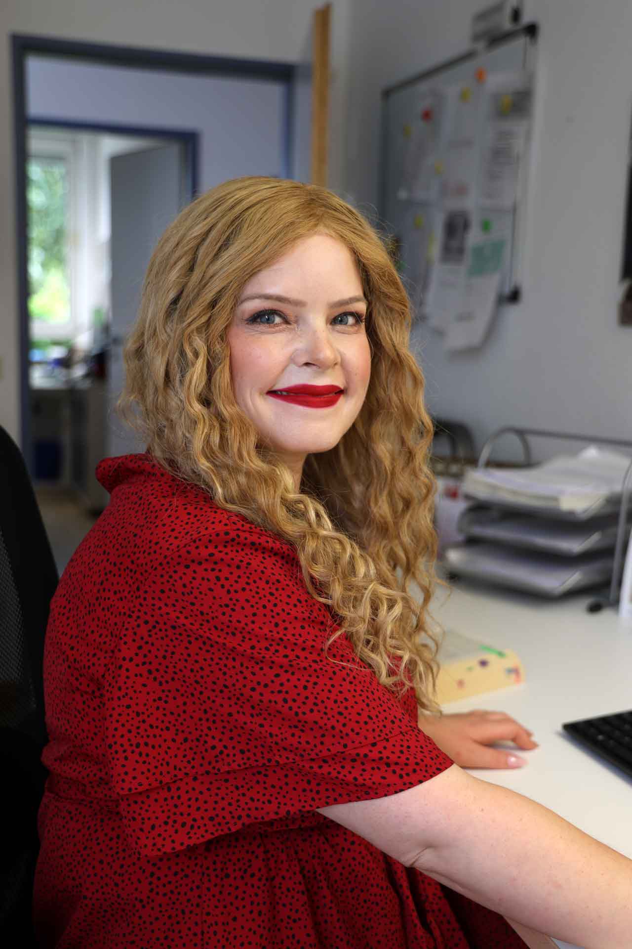 Juditz Desoi, eine Frau mit langen rotblonden Haaren. Sie trägt ein rotes Oberteil, roten Lippenstift und sitzt an ihrem Schreibtisch