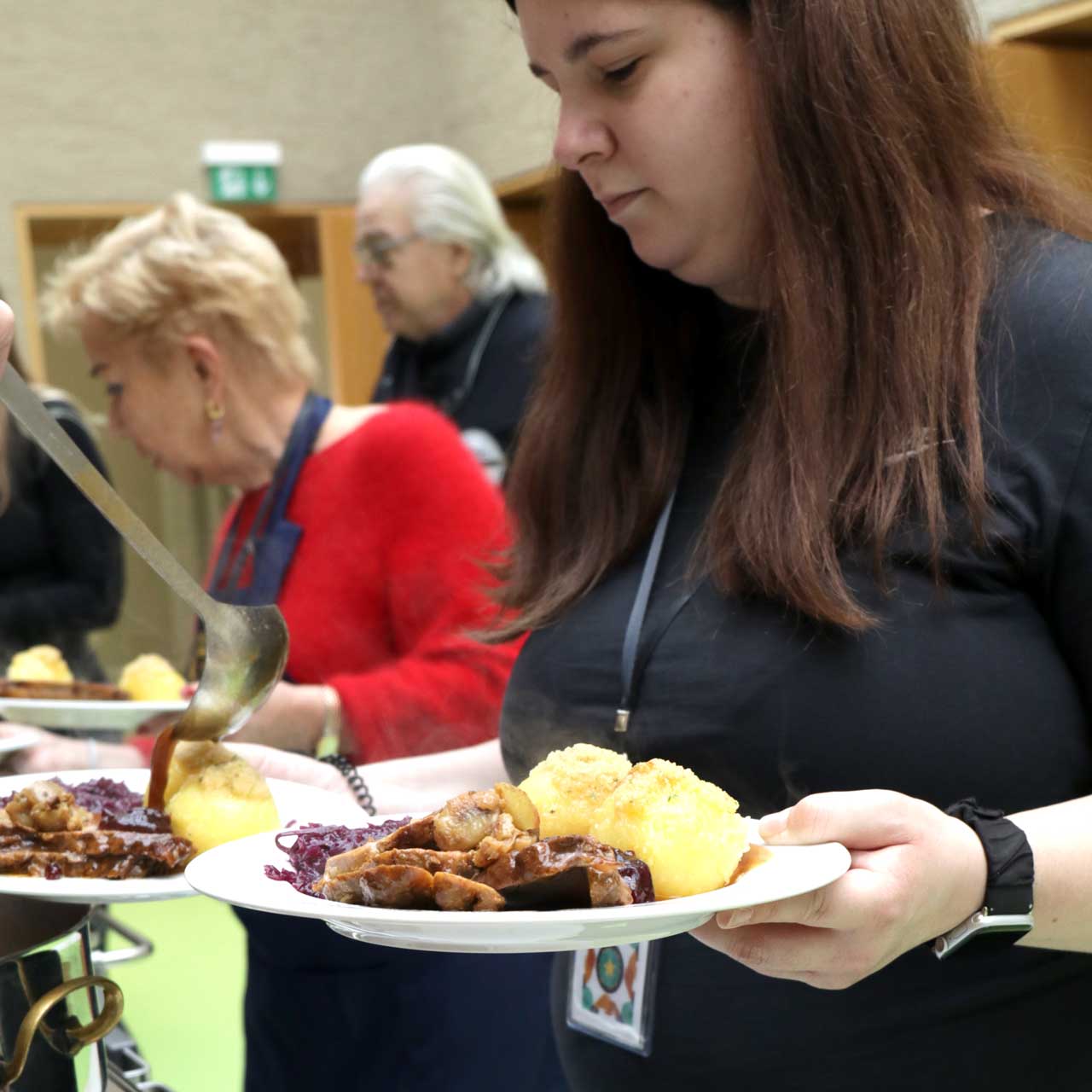 Eva hält zwei Teller mit dem Festessen in den Händen. Sie steht vor der Essensausgabe, wo ihr weitere Teile des Gerichts aufgetan werden.