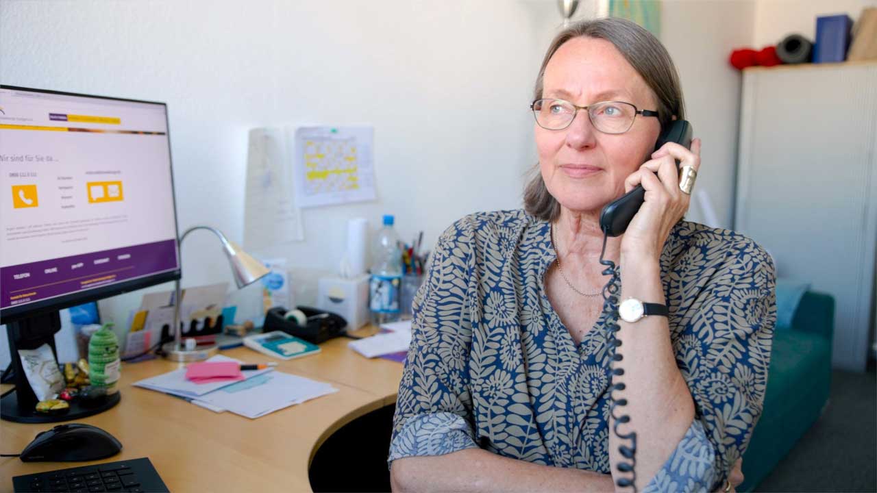Martina Rudolph-Zeller sitzt an ihrem Schreibtisch bei der Telefonseelsorge Stuttgart. Sie hält den Hörer an ihr Ohr und schaut nachdenklich. Im Hintergrund siehst du auf dem Bildschirm die Website der Telefonseelsorge. 