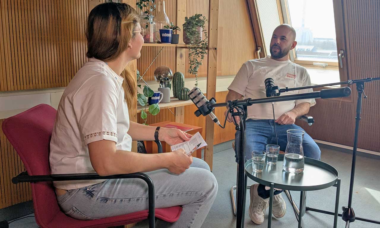 Host Charlotte Mattes, von hinten, von vorne sieht man sitzend Ricardo Sperlich. Mikros stehen im Raum.