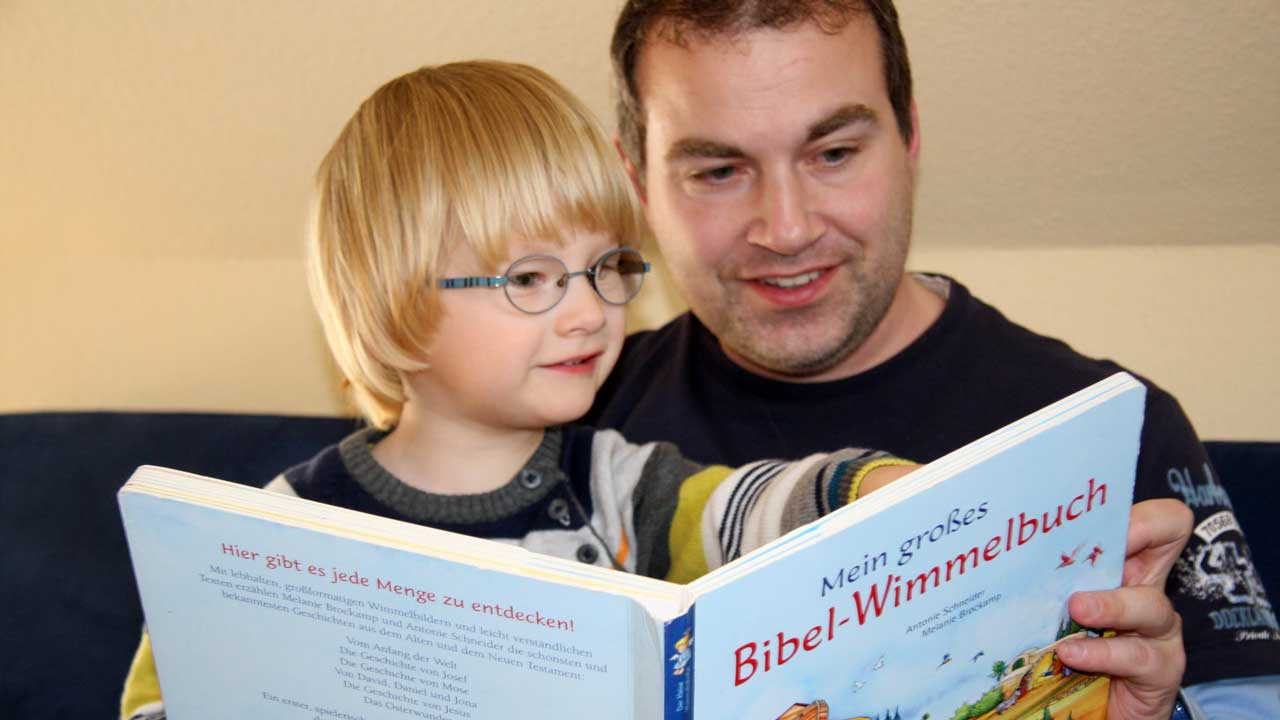 Gemeinsam mit dem Vater liest das Kind ein Bibel-Wimmelbuch.