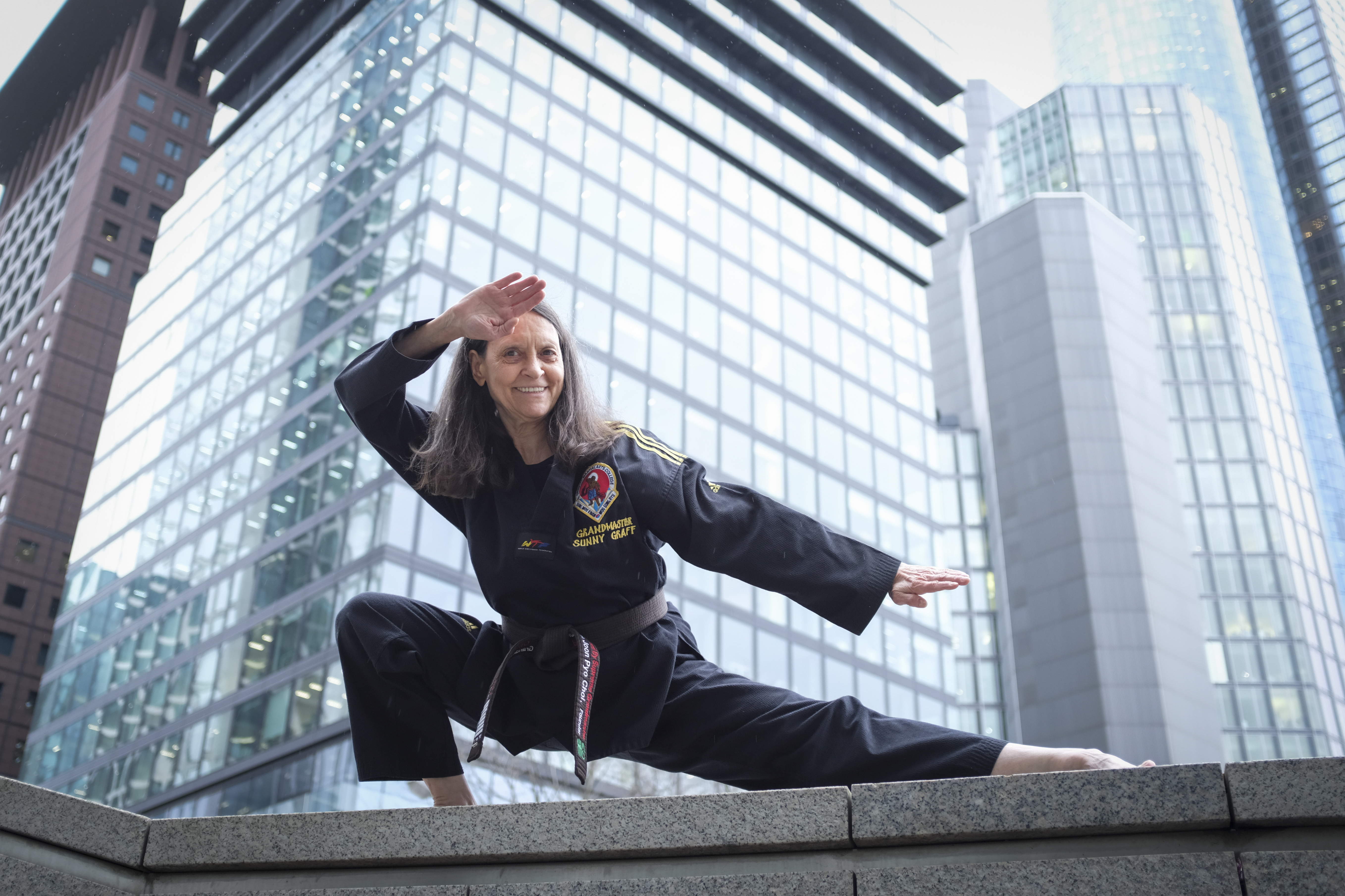 Die Takwondo-trainerin Sunny graff posioert in einer Taekwondo-position vor Hochhäusern, Sie trägt einen schwarzen Taekwondo-Anzug und lächelt.  