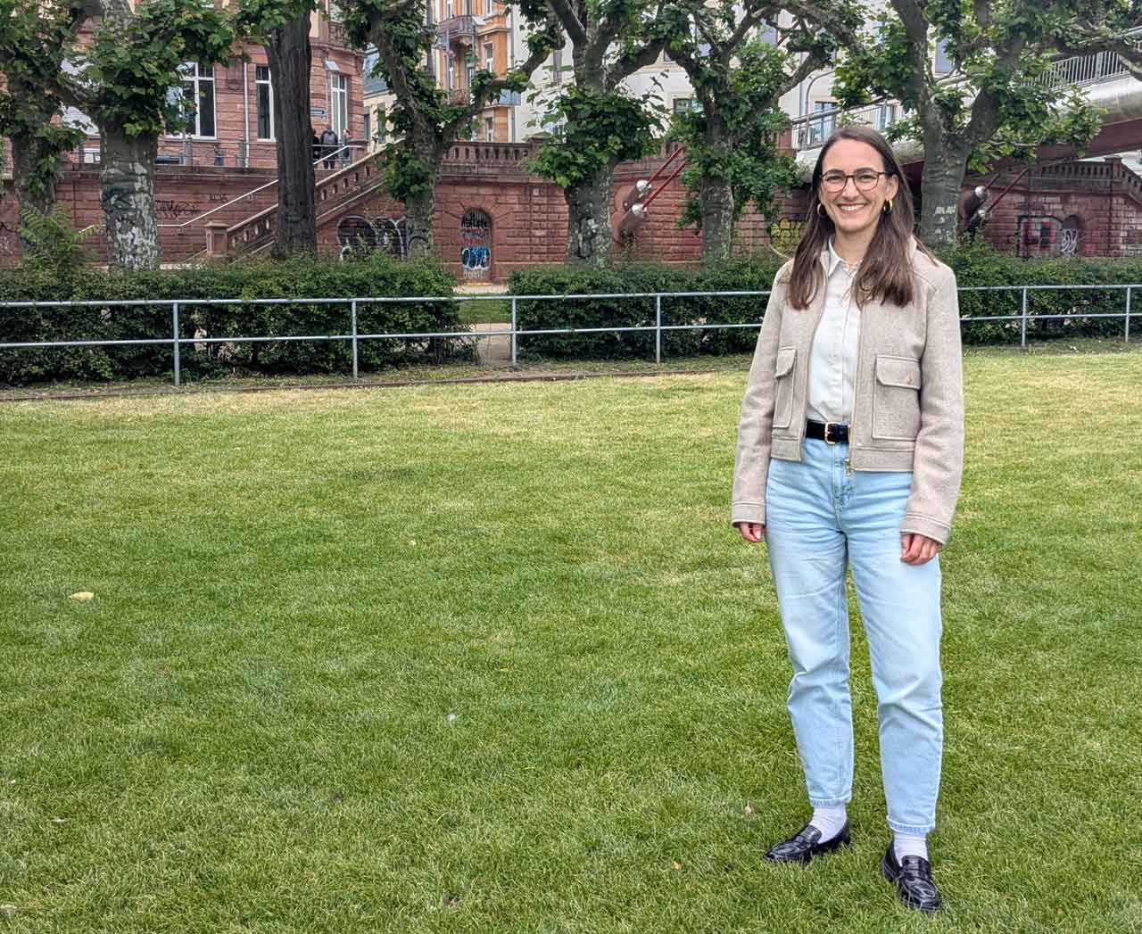Stephanie Hecke steht auf einer Wiese am Main, im Hintergrund sind Häuser und Bäume
