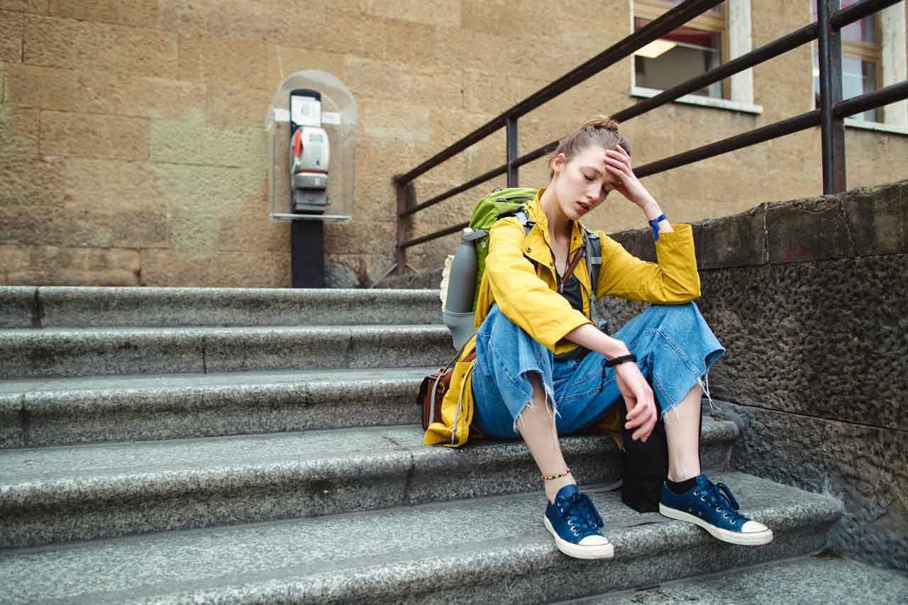 Das Bild zeigt eine junge Person, die mit Rucksack auf einer Steintreppe sitzt und sich erschöpft oder nachdenklich die Hand an die Stirn hält.