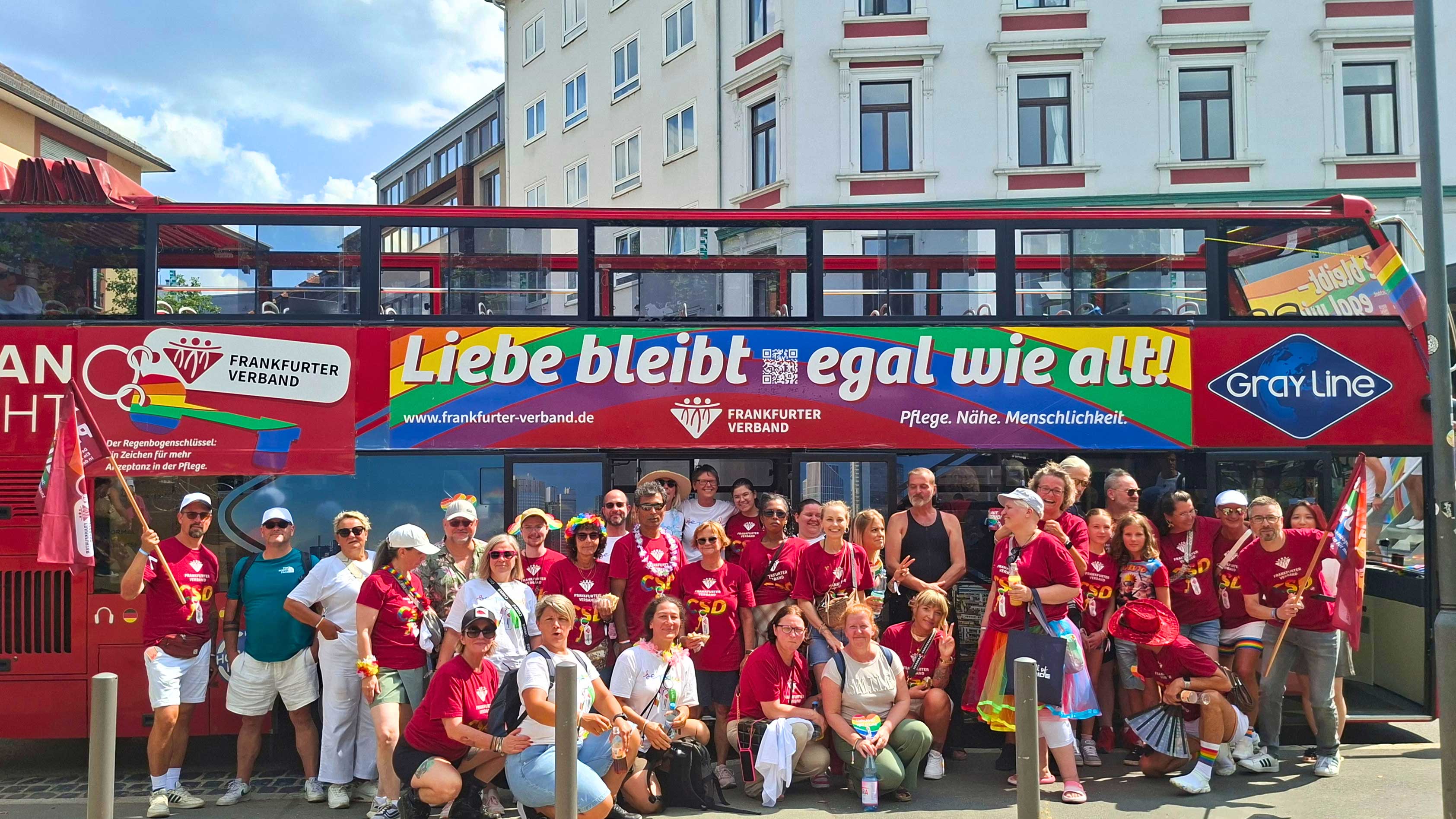 Eine Gruppe von Menschen steht vor einem roten Doppeldeckerbus mit Regenbogen-Design und dem Slogan „Liebe bleibt – egal wie alt!“. Viele tragen T-Shirts des Frankfurter Verbands und Regenbogen-Accessoires und posieren gemeinsam im Rahmen eines Christopher Street Day.