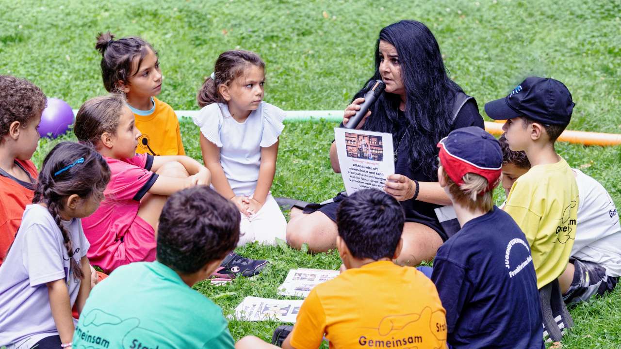 Raschida von Gemeinsam stark FFM sitzt im Kreis mit Kindern auf einer Wiese und klärt sie über Drogen auf