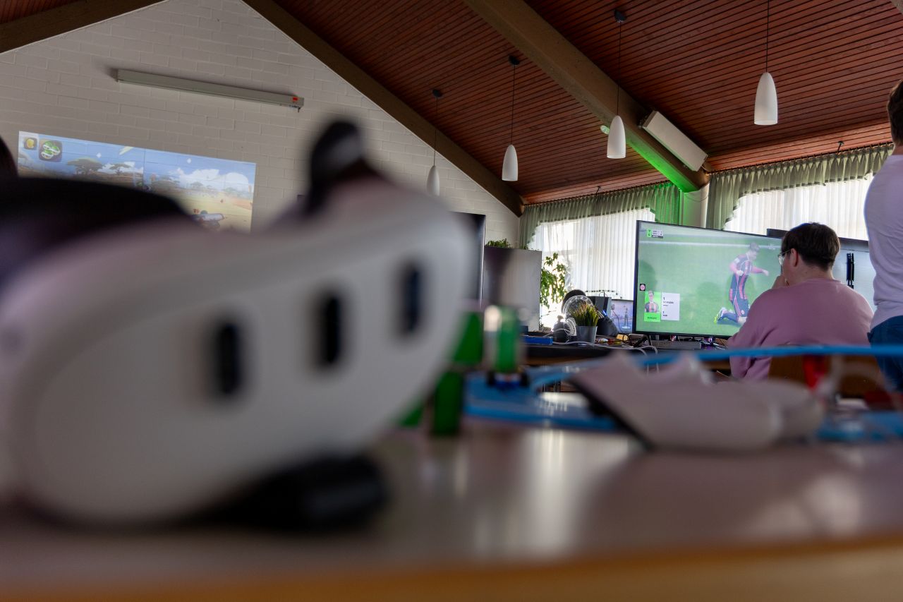 im Vordergrund liegt unscharf eine VR-Brille, im Hintergrund spielen Jugendliche Fifa auf einem großen Bildschirm