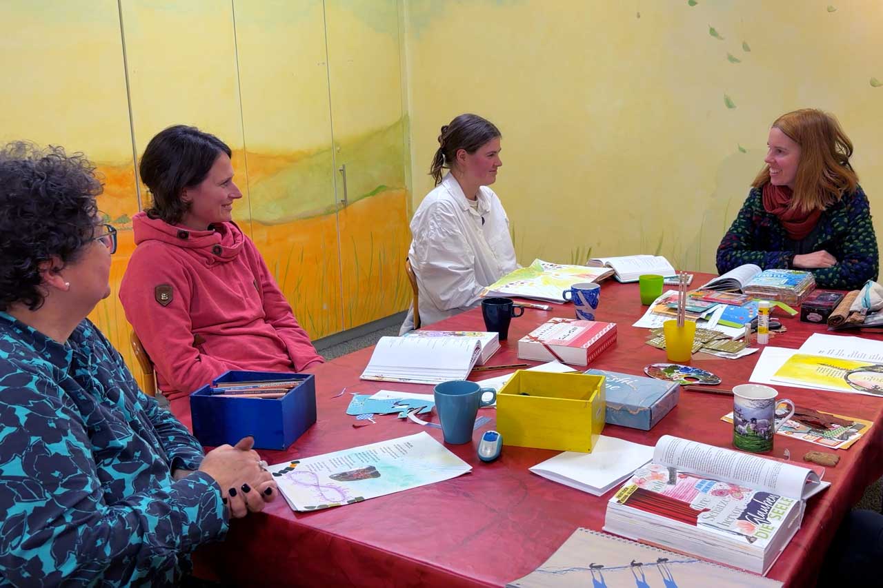 Eine Gruppe von Frauen sitzt um einen Tisch. Sie malen, schneiden und gestalten ihre Bibeln. Im Vordergrund sieht man eine Materialkiste. 