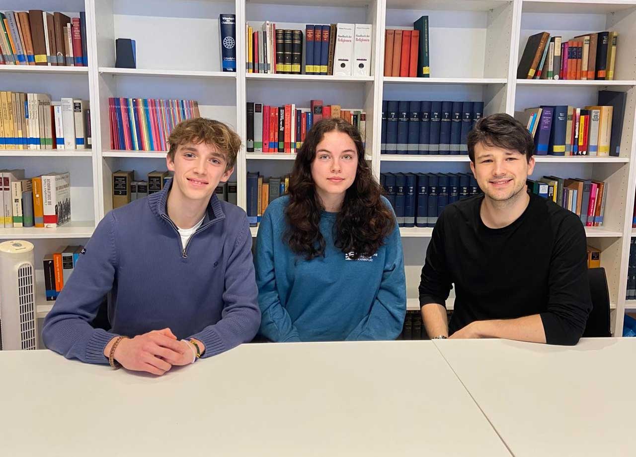 3 Personen sitzen vor einer Buchwand - in der Mitte sitzt eine junge Frau, rechts und links zwei junge Männer.