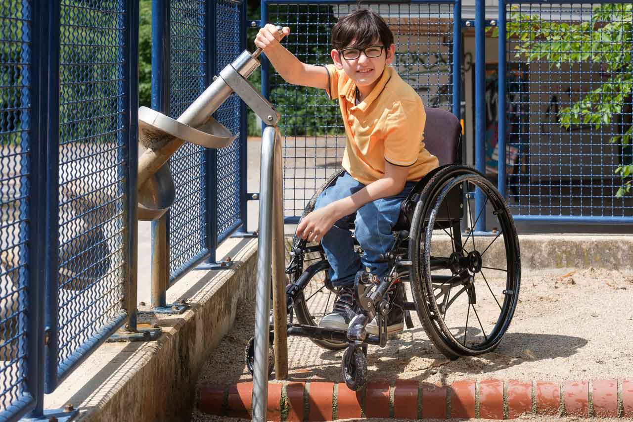 Metin sitzt im Rollstuhl und pumpf an einem der Wassergeräte Wasser. 